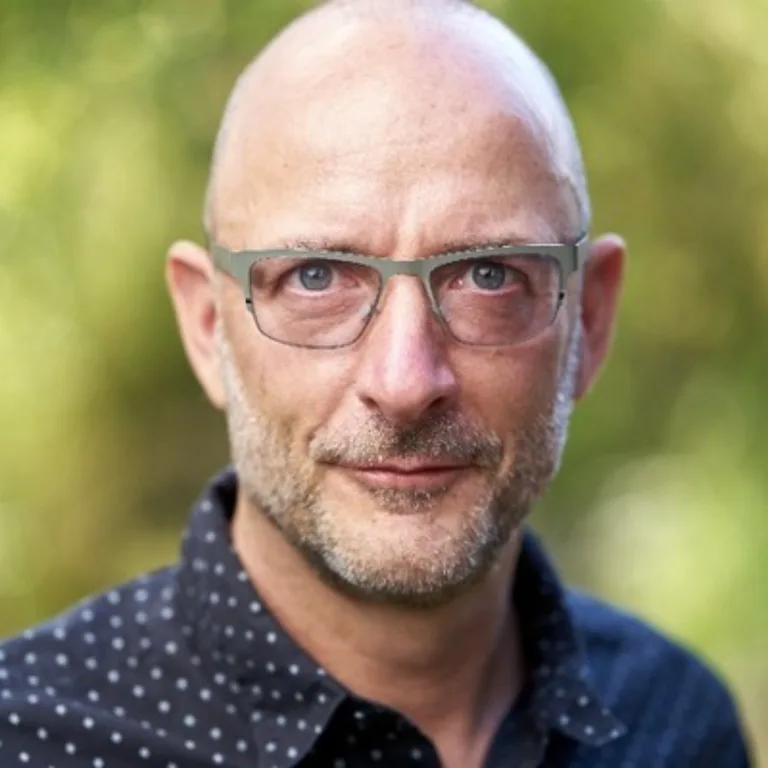 Mark Barden headshot, he is bald with a beard and wearing a blue and white polkadot shirt and glasses. He's standing in front of a green backdrop.