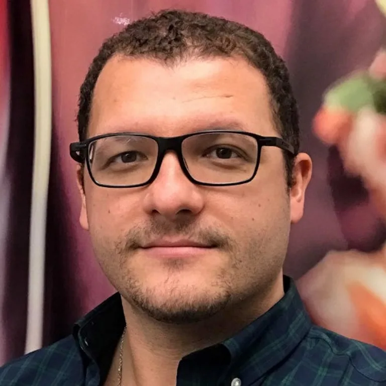 Walid Benchama headshot, he is wearing glasses and has short hair, standing against a red background.